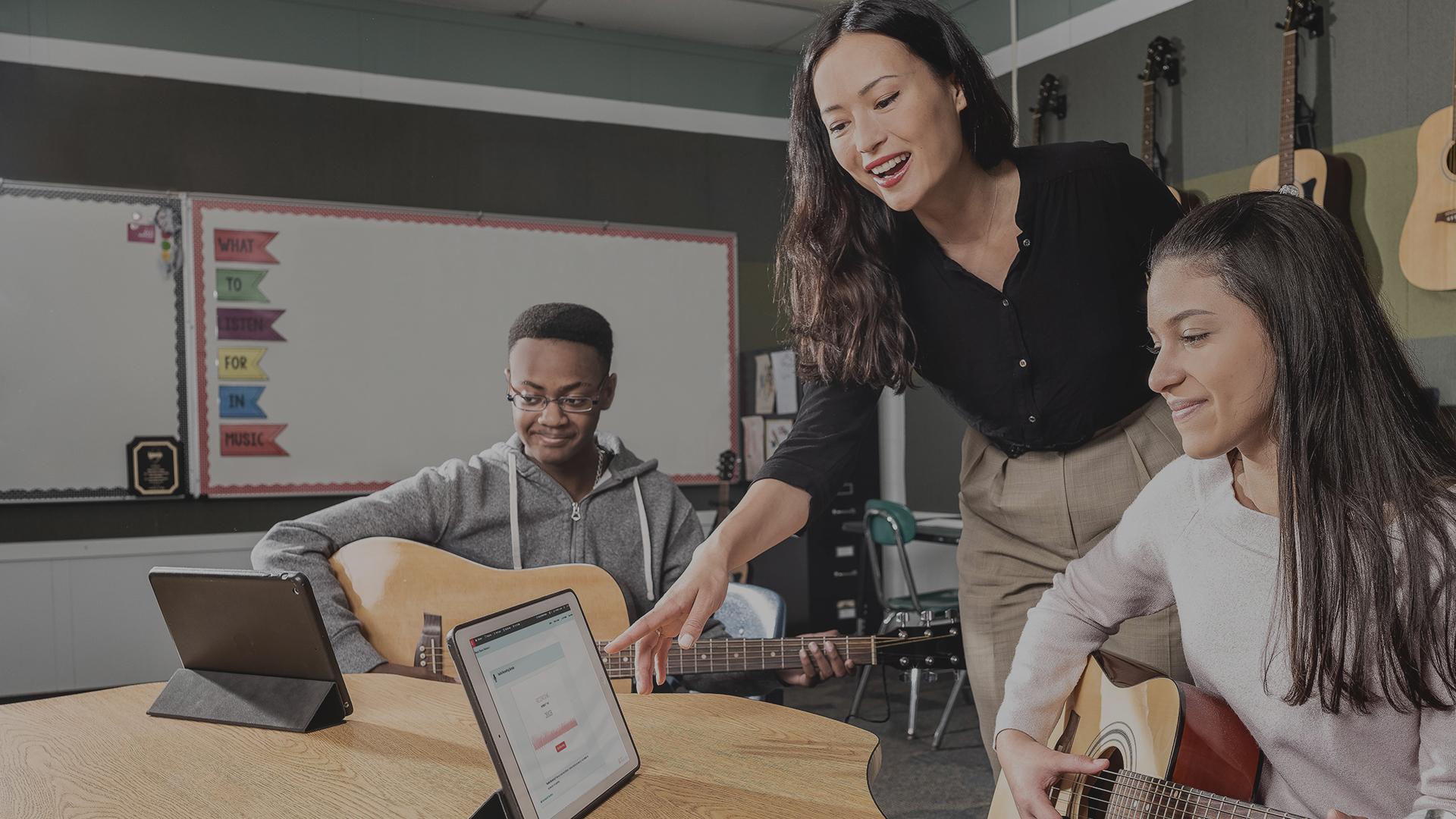 Teacher standing up pointing at a tablet. Two students sitting at a table holding acoustic guitars looking at tablets.