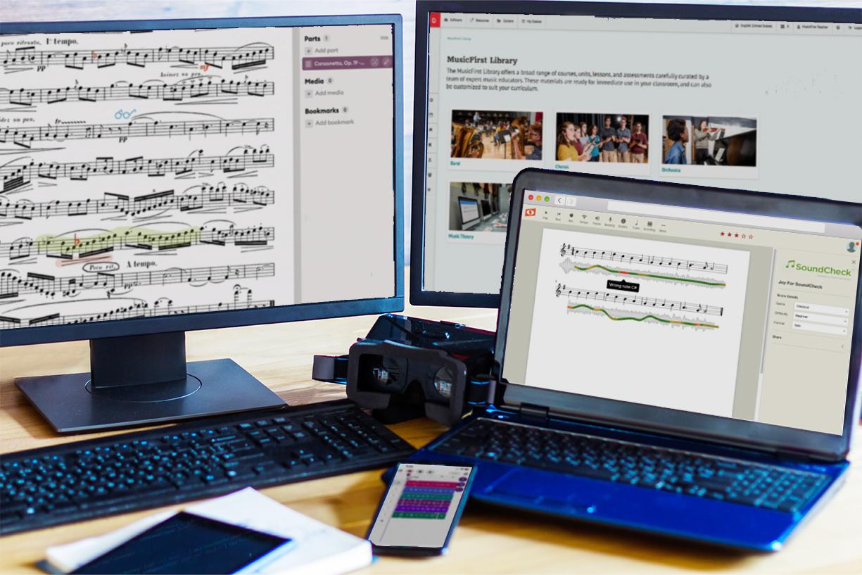 two desktop computers, one laptop, and a phone sitting on a desk. There is software open on each device (from left to right: Newzik Education, MusicFirst Classroom, Noteflight Learn, Soundtrap for Education)