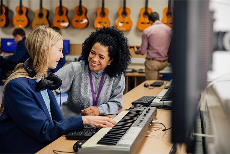 Student and teacher sitting together at a MIDI keyboard in front of a row of computers. There are acoustic guitars hanging on the wall in the background.