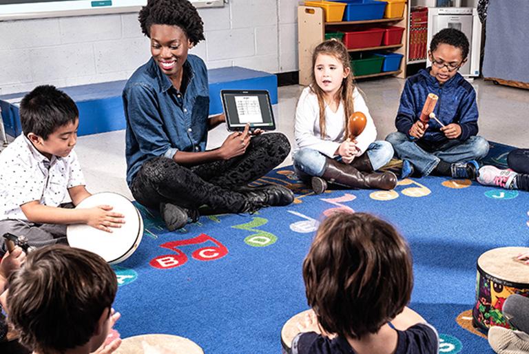 Teacher holder tablet sitting on a rug with young kids in a circle. Each of the kids is holding a musical instrument.