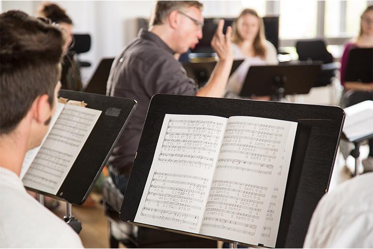 Over-the-shoulder of student standing at a music stand with sheet music on it, conductor in the background directing
