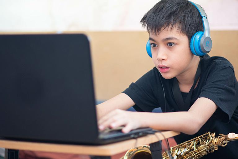 Kid wearing headphones sitting at a desk in front of a laptop. A saxophone is in his lap.