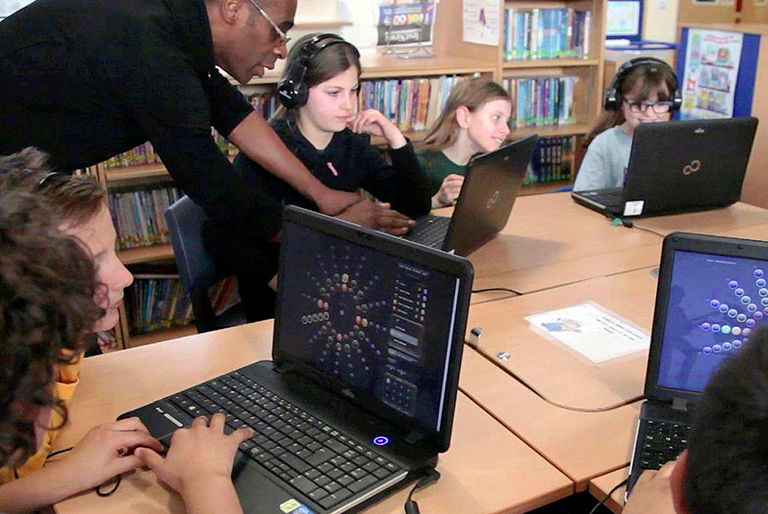 Students sitting at a table with O-Generator open on them. A teacher is assisting them with their composing