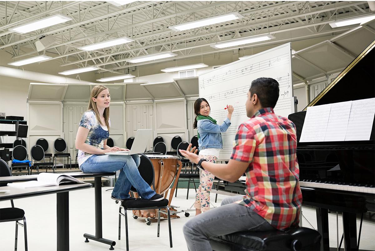 Three students sitting in a music rehearsal room. One student is sitting on a desk with a laptop in her lap, another is writing on a dry erase board, another is sitting at a grand piano.