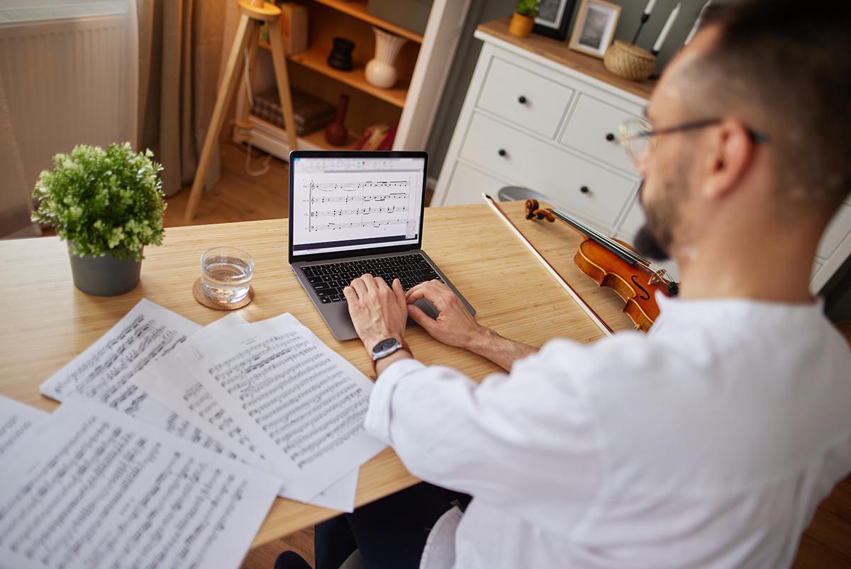 Man working on laptop editing sheet music. Paper sheet music is on the desk beside him.