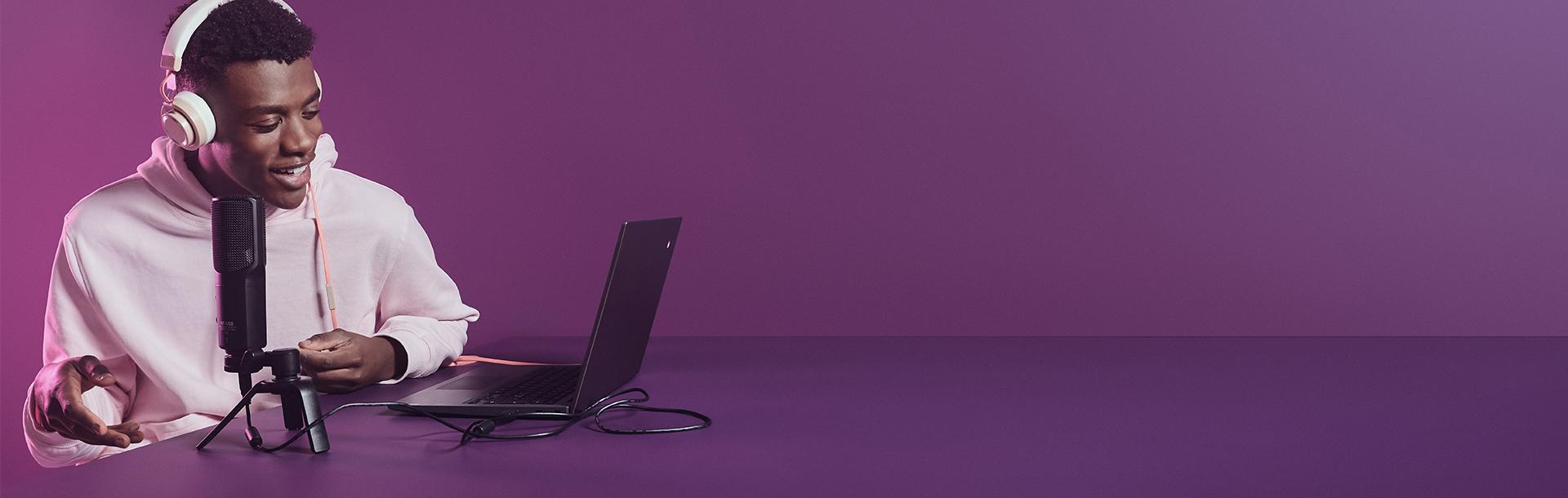Teenager in a white hoodie wearing headphones and recording into a microphone, looking at a laptop. The background is purple.